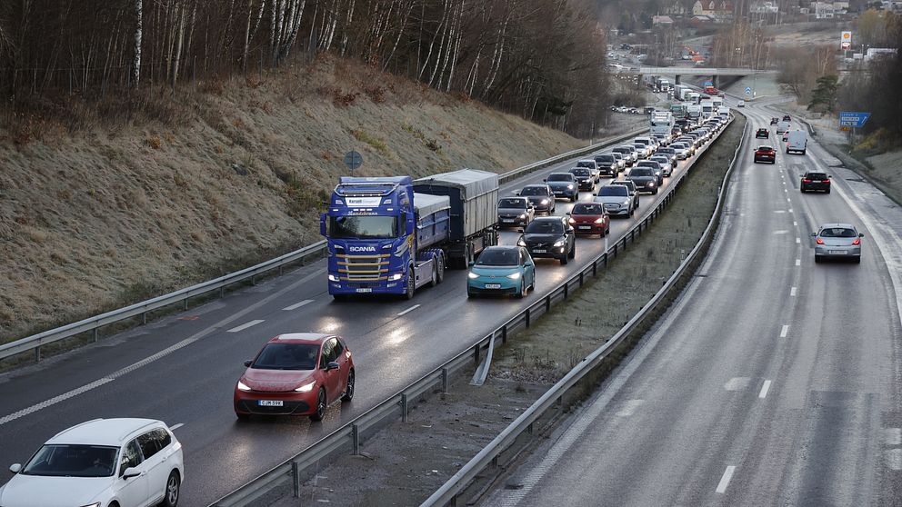 Kö av fordon på en motorväg, med en stor lastbil i fronten och flera personbilar som sitter fast i trafik. I bakgrunden syns skog och en bro, vilket indikerar en tät trafik och landskapsutsikt under en tidig morgon eller sen eftermiddag.