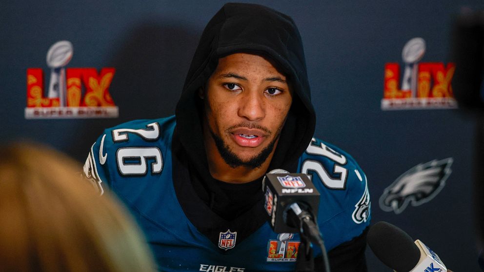 En amerikansk fotbollsspelare i Philadelphia Eagles-uniform, nummer 26, deltar i en presskonferens med flera mikrofoner framför sig. Eagles-logotyp syns i bakgrunden.