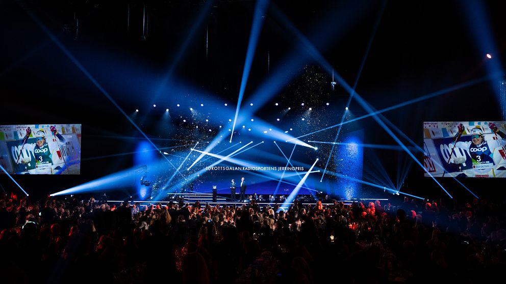 En scen i en stor evenemangsarena med starka ljusstrålar som strålar från taket. Publiken är samlad och delvis i mörker, med skärmar som visar bilder på scenen. Eventet verkar vara en prisutdelning eller gala, med diplomör och festlig atmosfär.