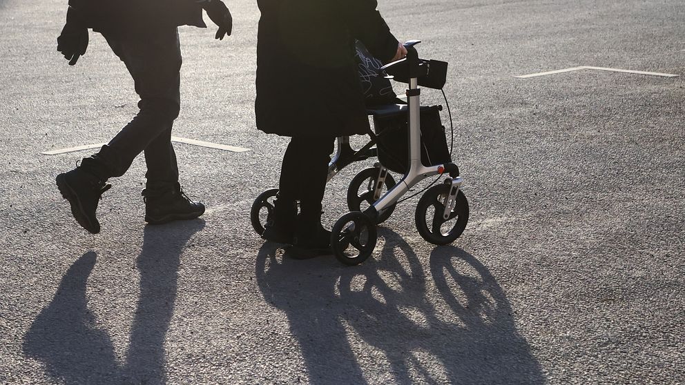 Halvbild på två äldre personer på en parkering. Mannen går själv och kvinnan har rullator.