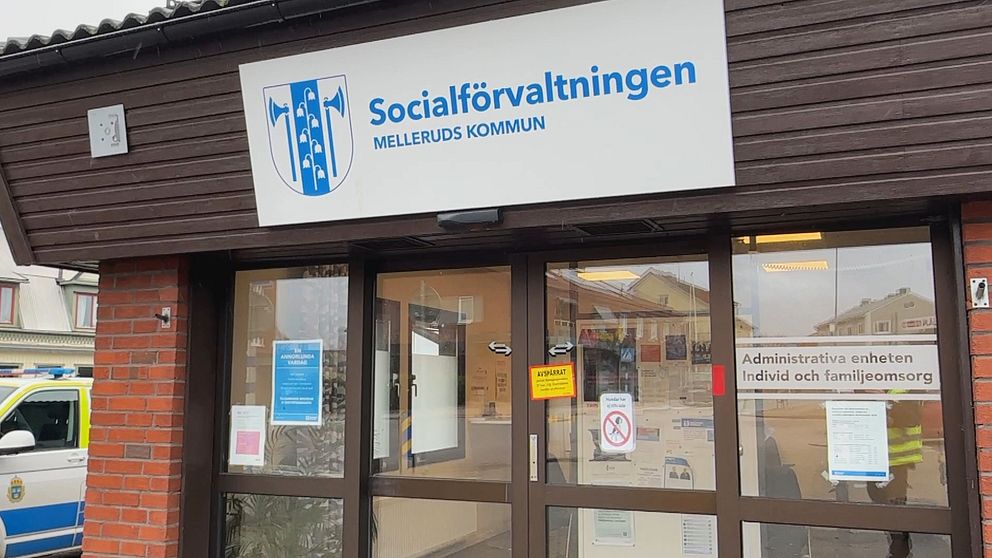 Entrén till Socialförvaltningen i Melleruds kommun, med en skylt som tydligt visar namnet. Flera informationsskyltar är synliga vid dörren, och en polisbil finns parkerad i bakgrunden.