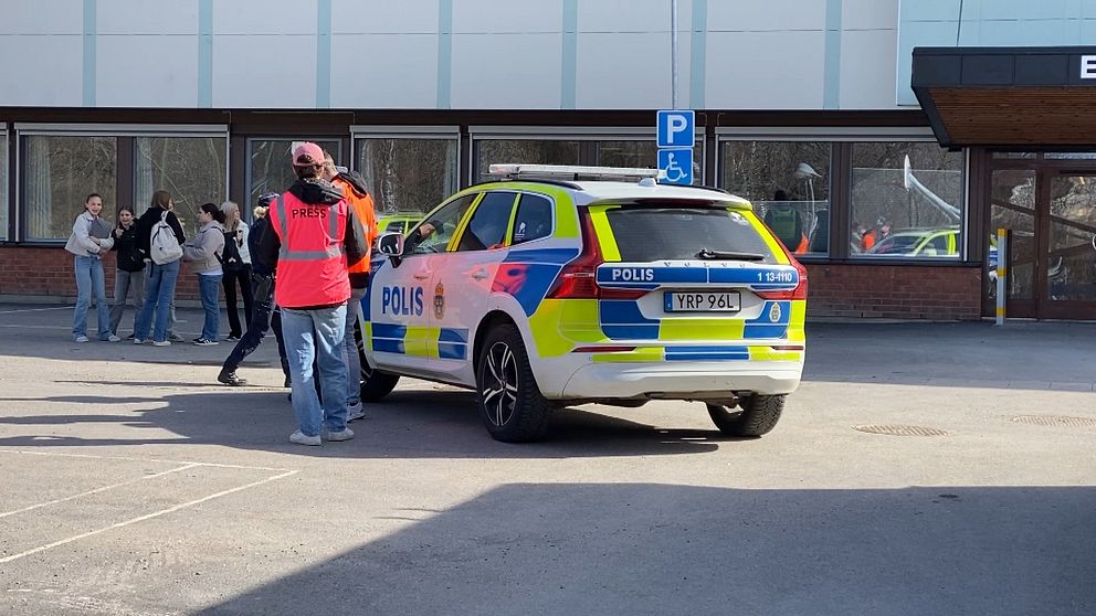 Polisbil parkerad utanför en byggnad. Person i röd väst pratar med flera människor.