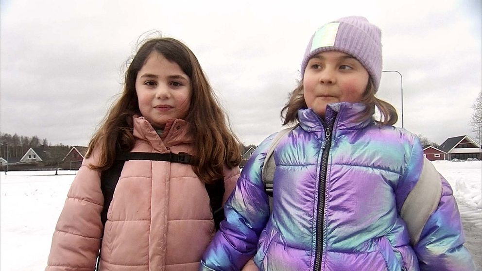 Två flickor står vända mot kameran intill en snötäckt idrottsplan