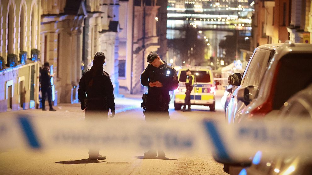 Poliser i uniform står mitt på en avspärrad gata i en stad under kvällen, med polisbilar i bakgrunden och stadsbelysningen som lyser upp byggnaderna i fjärran. En blå och vit avspärrningstejp syns i förgrunden.