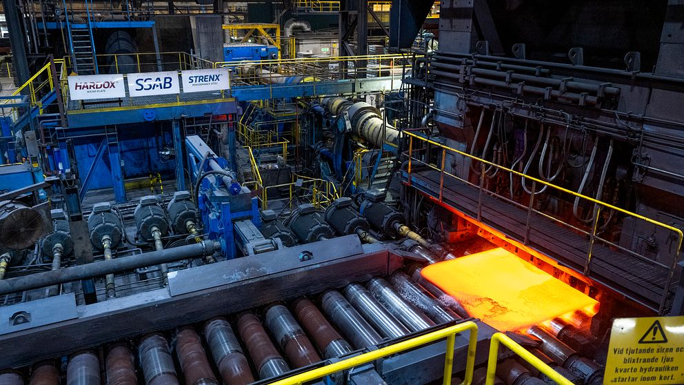 Rullande stålplatta i en industriell produktionslinje, omgiven av maskiner och utrustning. Plattan är lysande orange, vilket indikerar att den är uppvärmd. Skyltar med varumärken syns i bakgrunden tillsammans med en varningsskylt längst ner.