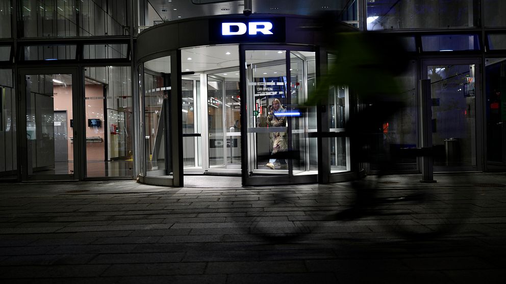 Entrén till DR:s byggnad i skymningen, oskarp cyklist i förgrunden.