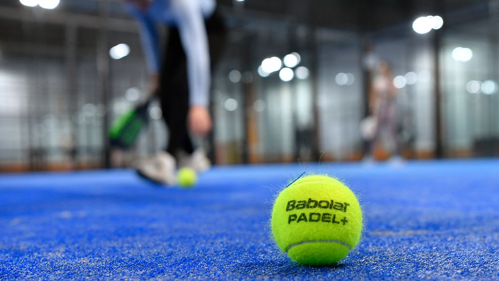 En padelboll ligger på ett padelbanans blå matta, suddig spelare i bakgrunden.