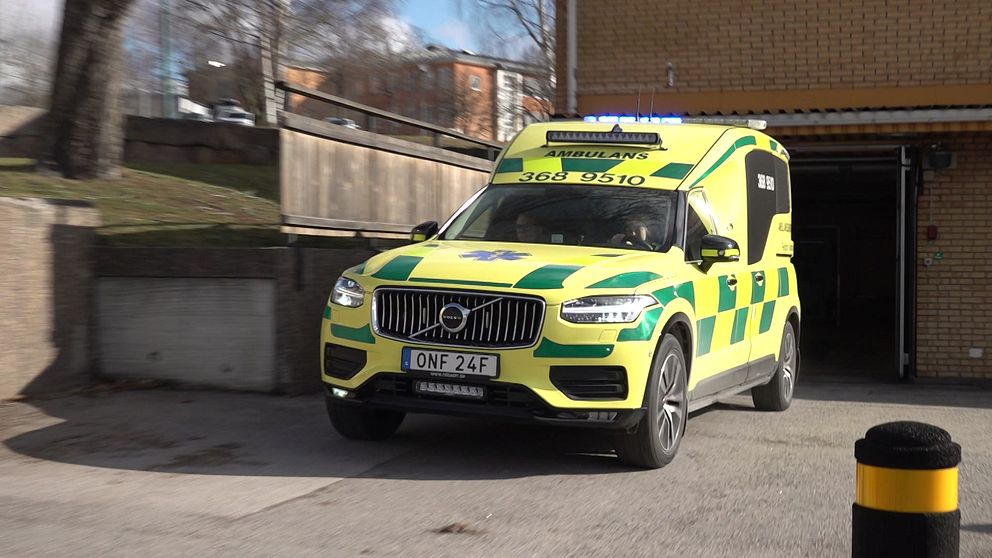 En ambulans i gul och grön färgsättning kör ut från en garageport i ett urbant område, med träddelar och byggnader i bakgrunden.
