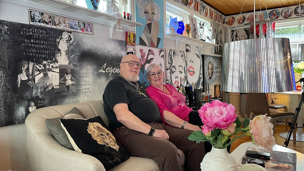 Ett par sitter på en beige soffa i ett inrett rum med en konstnärlig vägg prydd med bilder och citat. Alla bilder föreställer filmstjärnan Marilyn Monroe. Rummet har en modern taklampa och en blomma i en vas på bordet.