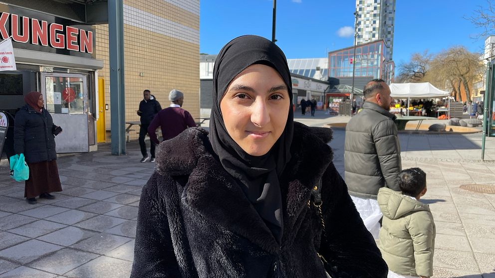 En ung kvinna står på Tensta torg i svart jacka och ler. Det går folk i bakgrunden.