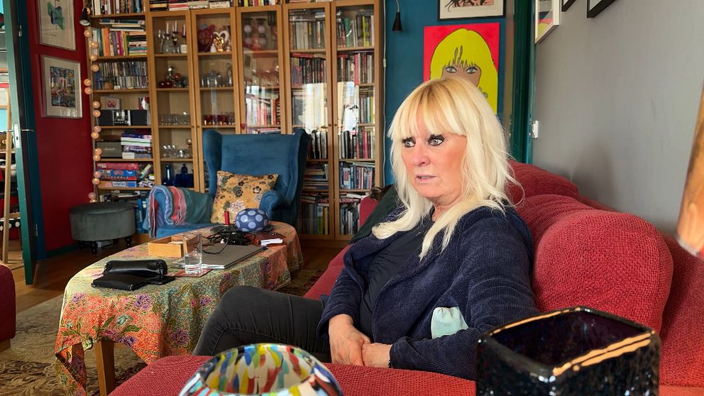 En kvinna sitter i ett rum fyllt med böcker och färgglada möbler, med en tavla synlig på väggen i bakgrunden. Miljön verkar personlig och hemtrevlig med olika objekt och en dekorativ duk på bordet.