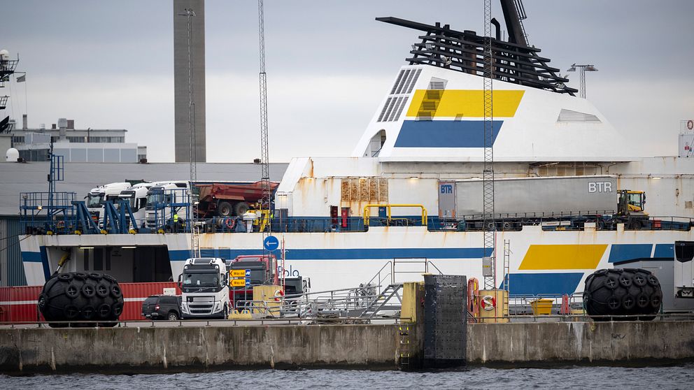 Färja vid kajen med vit och blå-gul randig överdel på ett varv, omgiven av industriell utrustning och lastbilar i en hamnmiljö.