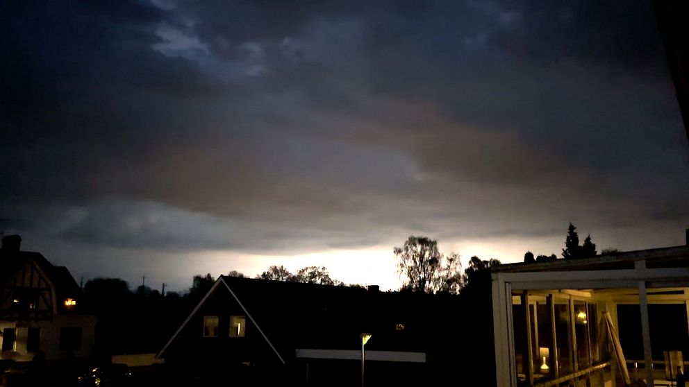 Mörka regnmoln med kraftiga blixtar över villaområde i skymning med siluetter av träd och hus mot horisonten.