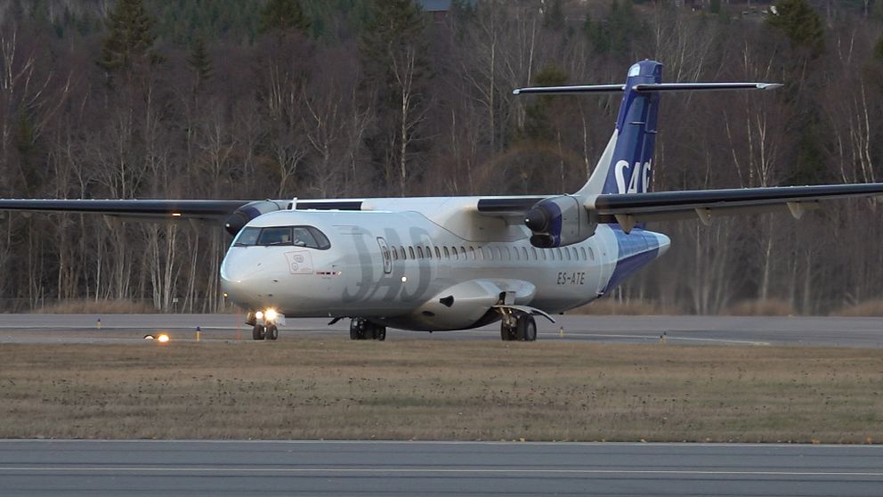 Ett SAS-flygplan taxar på en landningsbana omgiven av träd och en klar himmel. Flygplanet har en karaktäristisk vit och blå färgschema.
