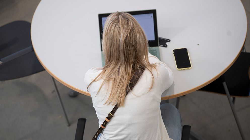 Kvinna, sedd bakifrån, jobbar vid laptop på rund bord med mobil bredvid.