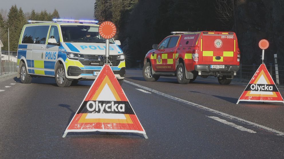 En polisbil och en räddningstjänstbil står på en väg som är avstängd med varningsskyltar som visar texten ”Olycka”. Varningsskyltarna är placerade på körbanan för att informera trafikanter om en incident. I bakgrunden syns träd och berg.