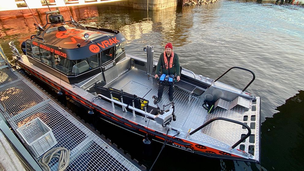 En ca 10 meter lång plåtbåt med styrhytt och stora öppna däck. Båten är uppmärkt med stora orange bokstäver ”VRAK”  – museet. PÅ däcket står en man med en undervattensdrönare i händerna. det ser ut som en liten u-båt.