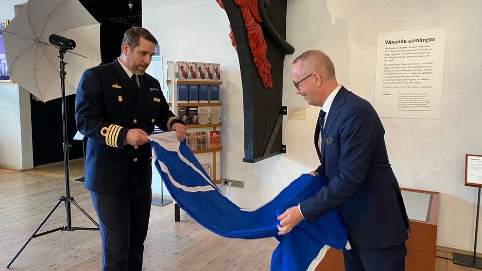 Två män i en formell miljö håller i en blå och vit flagga. Ena mannen är klädd i en uniform och den andra i kostym. De står bredvid en utställningsplats med en skylt i bakgrunden som säger ”Utställningar”.