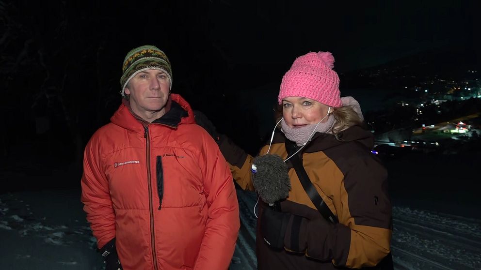 En person i röd jacka står bredvid en person i mörkare kläder och en rosa mössa, medan de pratar i ett snölandskap under natten. Staden syns svagt i bakgrunden, belyst av ljus.