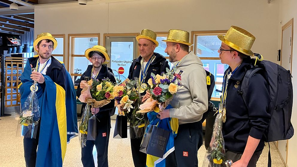 Fem personer med guldhattar står i grupp och håller blommor.