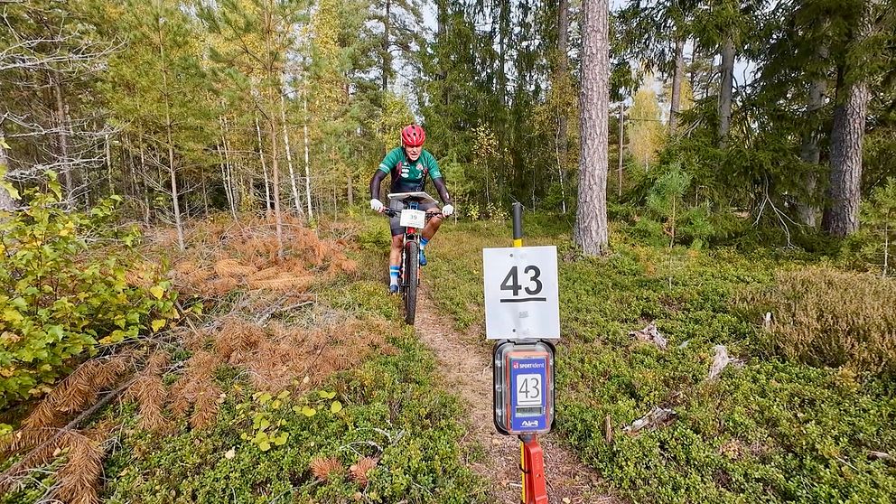 En cyklist på en stig i en skog passerar en skylt med nummer 43. Omgivningen består av grönskande buskar och träd, med höstliknande färger i markvegetationen.