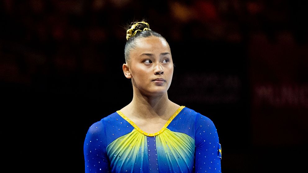 En gymnast i en färgglad, glittrande dräkt med blå och gula detaljer står på scenen under en tävling. Bakgrunden visar en oskarp åskådararena, vilket ger en känsla av en spännande sporthändelse.