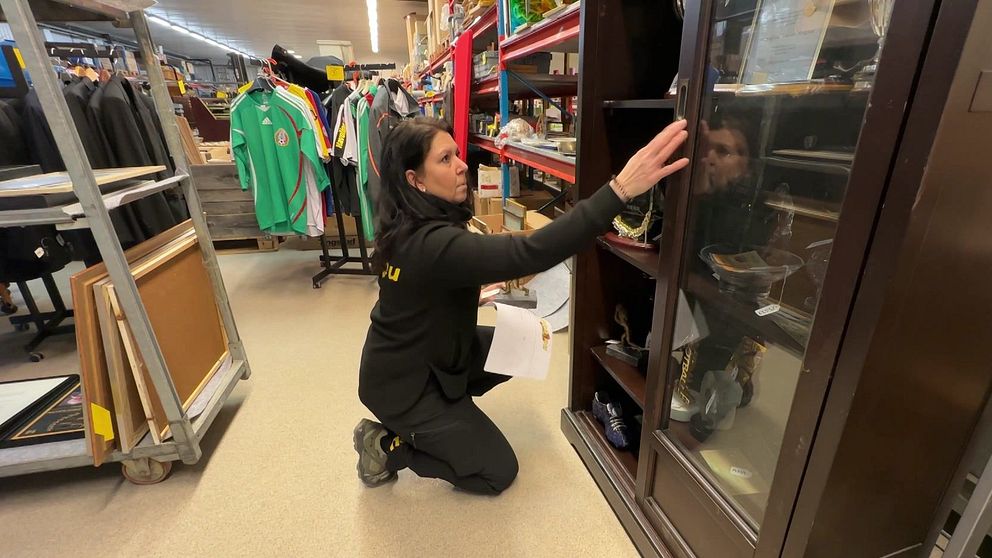 Kvinna i lokal organiserar hyllor med glasdörrar, omgiven av kläder och varor från auktionen av Svennis kvarlåtenskap.