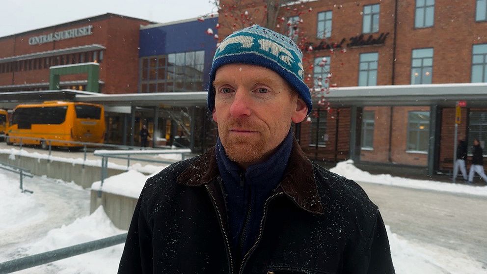 En person står framför Karlstad centralsjukhus med flera fönster och snö på marken. I bakgrunden syns en gul buss och en del av en annan byggnad med en skylt. Det är vinter och snöigt.