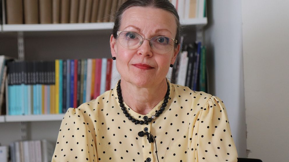 Birgitta Meurling i gul prickig blus sitter framför bokhylla.