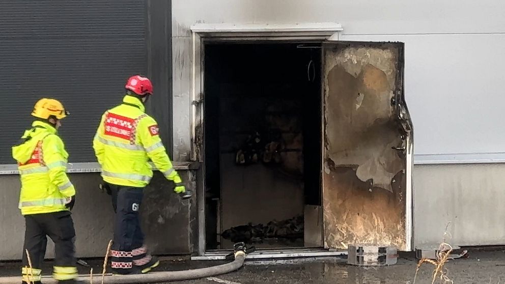 Två brandmän i gul och svart skyddsutrustning står vid en brinnande dörröppning till en byggnad. Dörren är delvis öppen och visar tecken på brandskador, med svart sot på väggarna inuti. En brandslang ligger på marken framför dem, vilket indikerar pågående släckningsarbete.