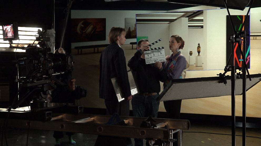 En filmproduktion pågår inomhus, där en person står framför kameran och förbereder sig för en scen. En annan person håller en klappa (slate) medan två personer diskuterar i bakgrunden. I rummet syns en konstutställning med olika verk på väggarna, vilket ger en avslappnad och kreativ atmosfär. Kamerautrustning syns vid sidan och belysning används för att belysa scenen.
