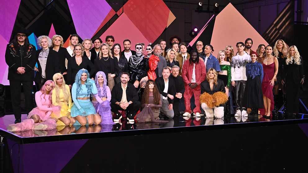 Gruppbild av deltagare och artister från Melodifestivalen 2025, klädda i färgglada och varierande kläder, poserande på en scen med geometriska bakgrundselement.