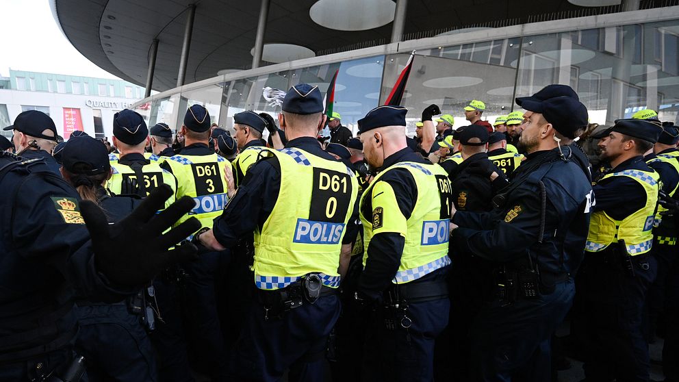 Grupp av poliser klädda i gula västar med ”POLIS” tryckt på framsidan, samlade utanför en byggnad. De är omgivna av en stadsbild och verkar vara inblandade i en säkerhetsinsats eller ett offentligt evenemang.