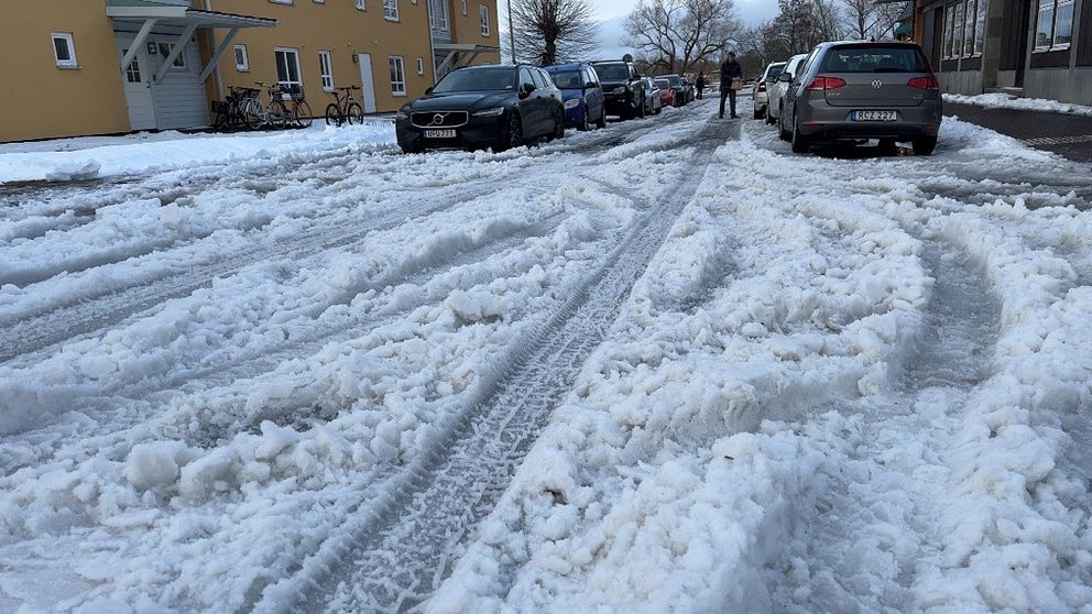 En snötäckt gata med djupt snö spår efter bilar. Parkerade bilar syns på båda sidor om vägen, och en person går på den snöiga trottoaren.