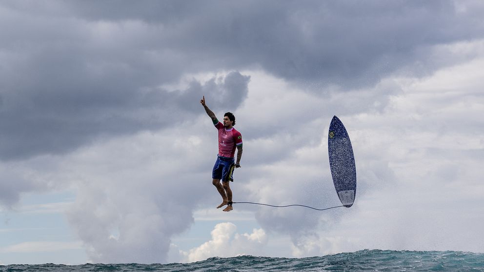 Surfare i rosa tröja hoppar över vattnet med en surfbräda i handen, omgiven av mörka moln och en stormig himmel.