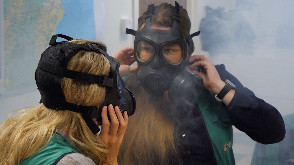 Två personer står i ett rum och justerar sina gasmasker medan röken sprider sig kring dem. Båda bär gröna västar och syns i en utbildnings- eller träningstillställning som handlar om säkerhet eller krisberedskap. En karta kan ses i bakgrunden, vilket tyder på att platsen är inomhus.