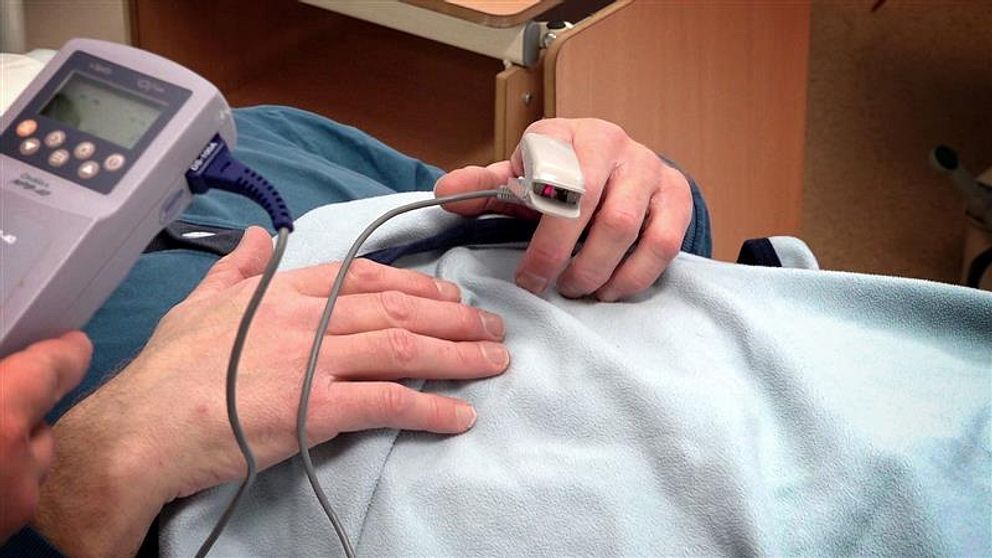En person i sjukhussäng mäter syremättnad med pulsoximeter på fingret.
