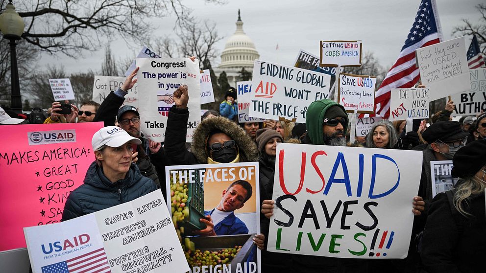En grupp människor står samlade framför Capitolium i Washington D.C. och håller upp skyltar som stöder USAID, där budskap som ”USAID SAVES LIVES” och ”THE FACES OF FOREIGN AID” är framträdande. Många deltagare håller också amerikanska flaggor, och stämningen i bilden tyder på en aktiv demonstration för biståndsmyndigheten.