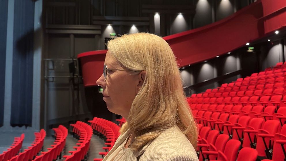 En person med långt blont hår står i profil i en teater. I bakgrunden syns ett stort antal röda stolar i publikområdet, med en mörk scenyta till vänster.