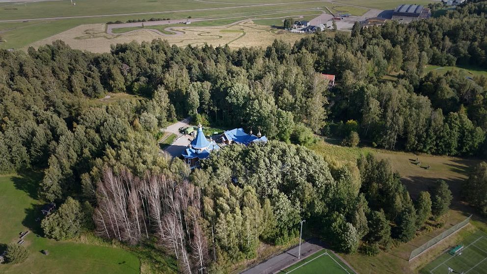 Flygfoto över ett hus med blått tak omgivet av tät skog och öppna fält.