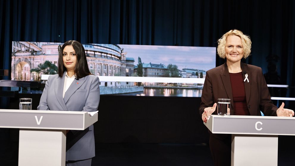 Två politiker står vid talarpodium under en debatt med riksdagsbakgrund på skärmen.
