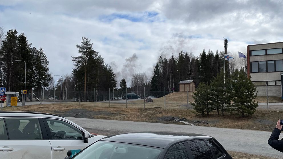 Rök stiger bakom ett staket nära en skola eller industribyggnad, bilar i förgrunden.