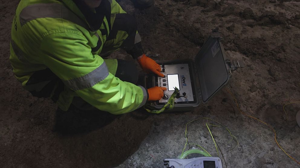 En person i skyddskläder med lysande gul jacka sitter på huk på en jordig mark och hanterar en teknisk enhet i en portabel låda, omgiven av sladdar och en elektronisk mätinstrument.
