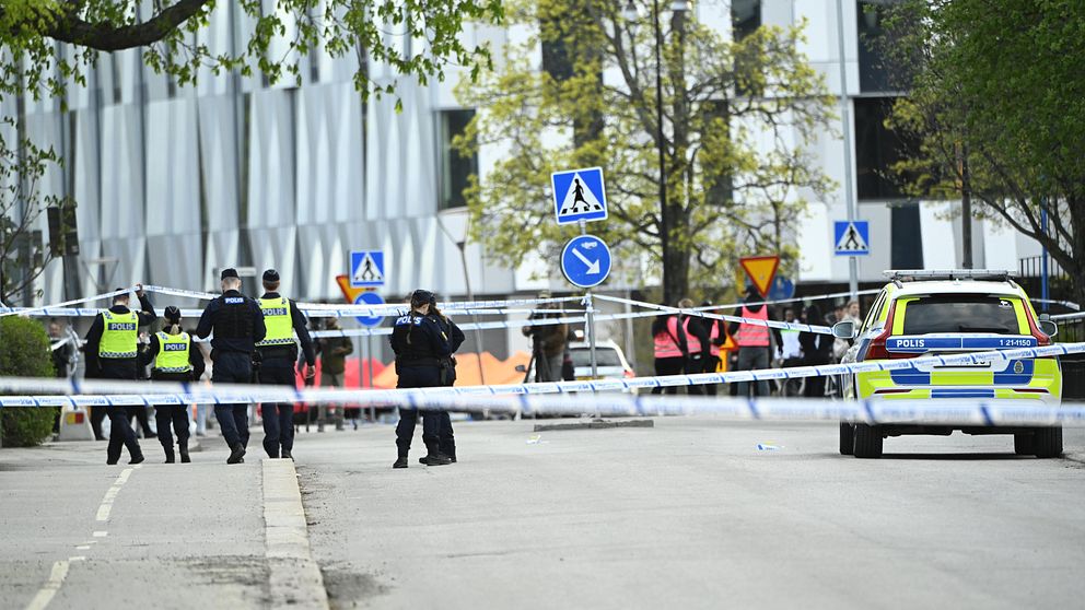 Poliser vid avspärrning och polisbil på gata utanför en modern byggnad.