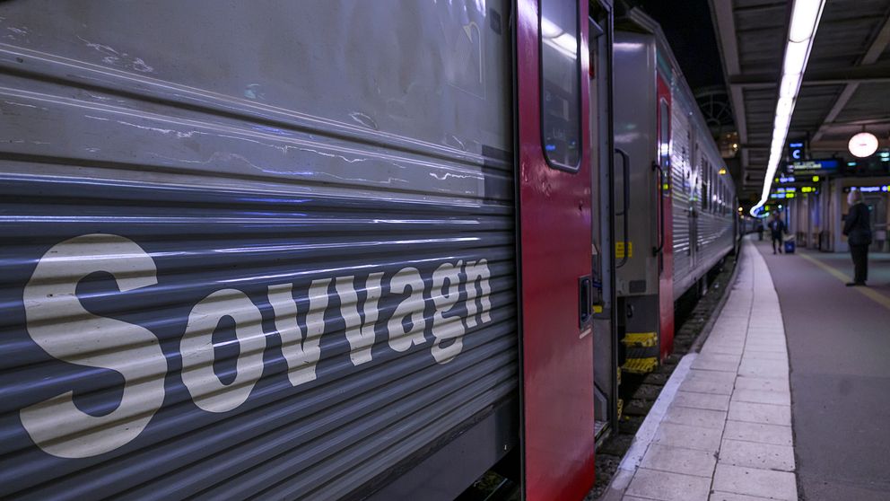 ALT-text: Tåg med texten ”Sovvagn” på sidan, stående vid en perrong i mörker, med plattformsbelysning och skymtor av människor som väntar.