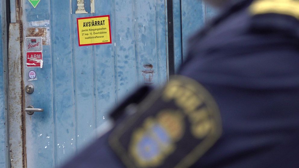 En ytterdörr med en gul skylt som varnar att området är avspärrat. I förgrunden syns en del av en polisuniform med emblem, vilket indikerar att polis är närvarande vid platsen.