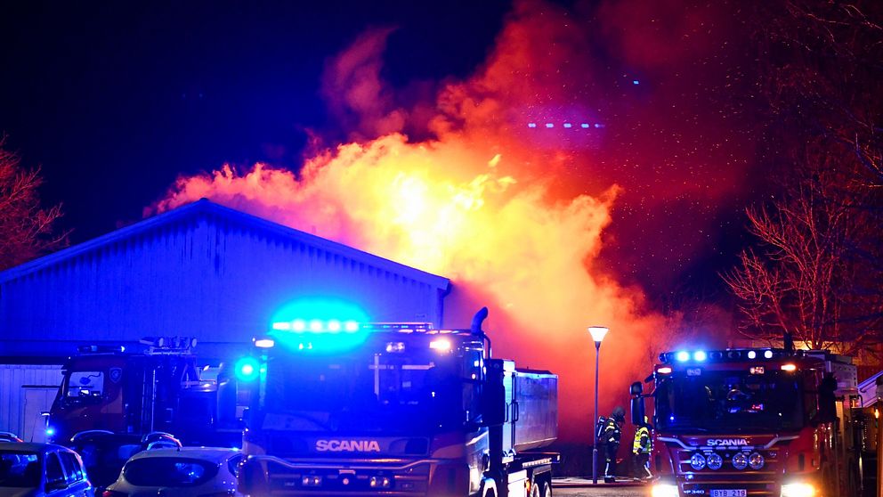 Ett brandutryckningsscenario med flera brandbilar vid en kraftigt flammande byggnad i Vara. Blåljusen från räddningstjänsten lyser upp omgivningen.