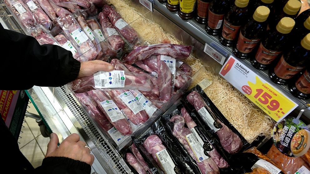 En person väljer bland olika sorters kött som ligger på ett kylskåp i en butik. I bakgrunden syns flaskor med sås och prisinformation för köttet.