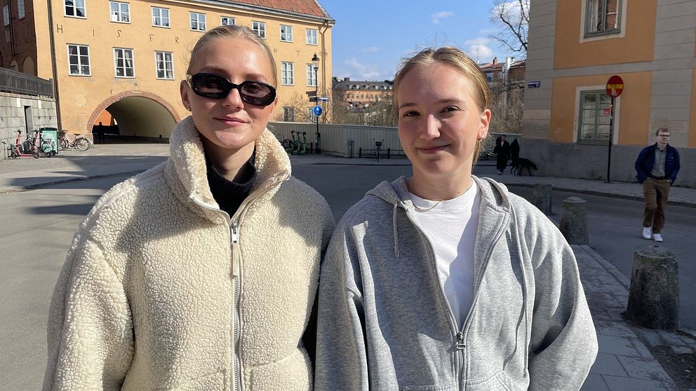 Två personer står utomhus i en solig stadsmiljö. Gamla byggnader i bakgrunden.