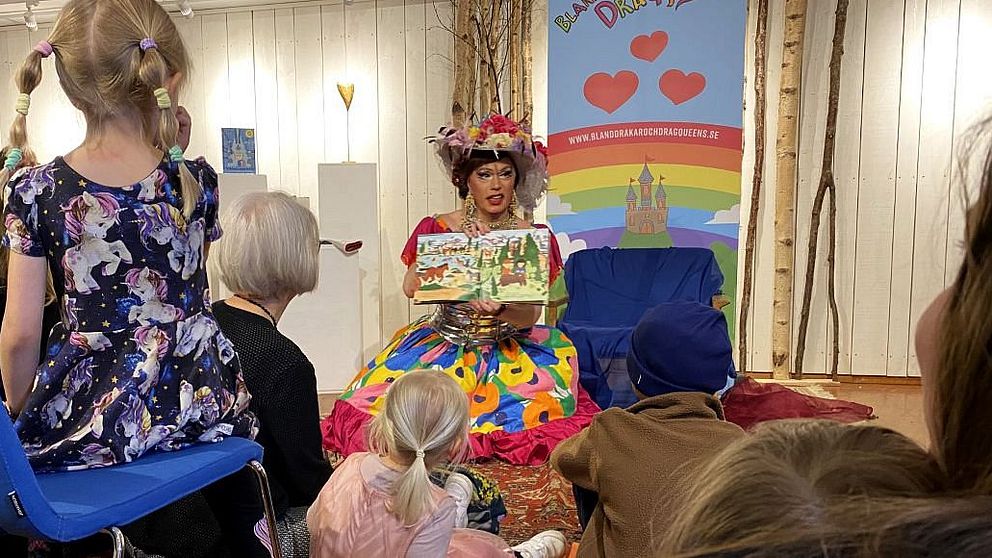 Person klädd i färggrann klänning läser bok för barn på bibliotek. Barnen sitter på golvet.